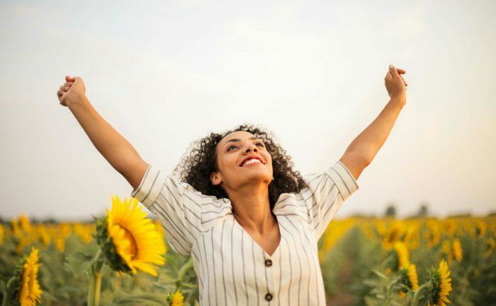 A joyful woman standing in a sunflower field with her arms raised in the air, smiling brightly under a clear sky.