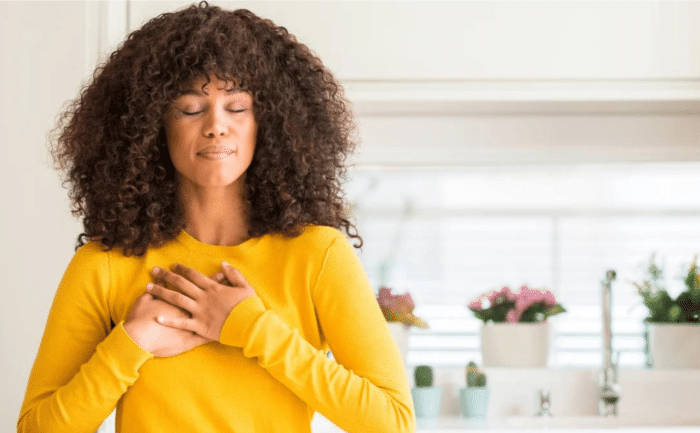 Woman practicing gratitude with her hands over her heart, eyes closed, wearing a yellow sweater in a bright room with plants in the background.