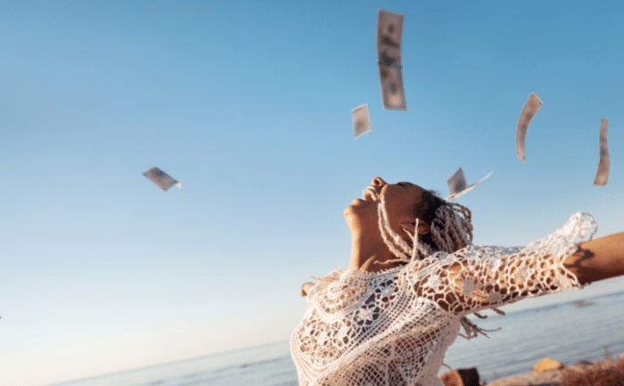 A joyful woman with braided hair and a white crochet top is standing by the sea, her arms outstretched, as currency notes float around her in the air.