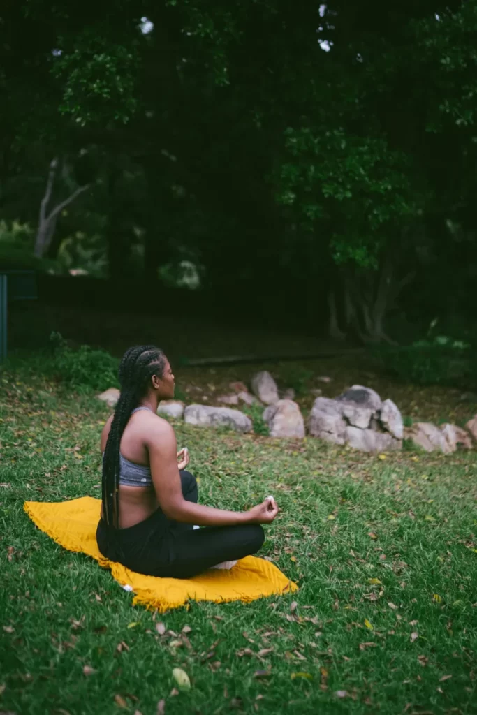 A woman sits in a peaceful outdoor setting, meditating on a yellow mat surrounded by greenery.