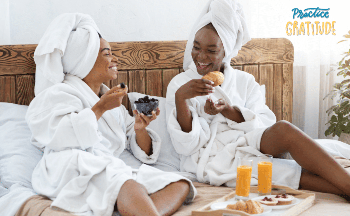 Two women in white robes and towels on their heads enjoy a healthy breakfast in bed, laughing and sharing a moment of joy. They are eating fruits and pastries, with glasses of orange juice on the tray beside them, in a bright, cozy bedroom setting.