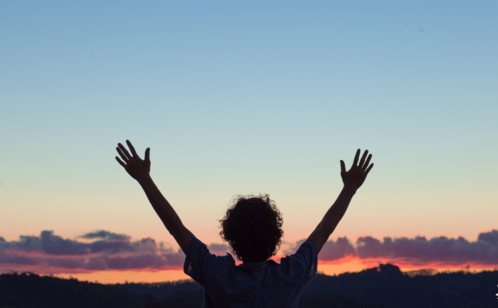 A silhouette of a person with curly hair, arms raised in celebration against a vibrant sunset sky. The scene conveys a sense of freedom and gratitude.